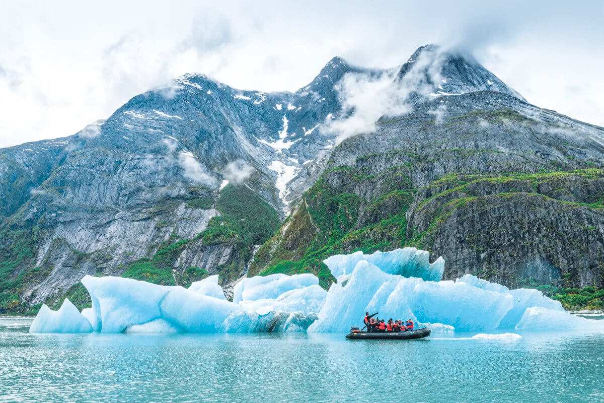 A passenger-filled zodiac passes by an ice-blue iceberg in Misty Fjords